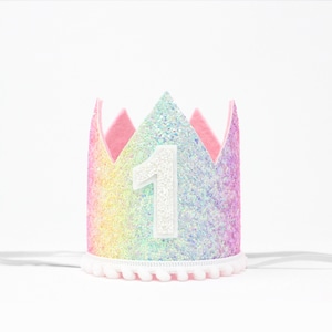 May include: A crown featuring a rainbow-colored glitter finish with a white glitter number one centered on the front. The crown has a pink felt interior lining, a white pom-pom trim along the bottom edge, and thin white elastic straps on each side.