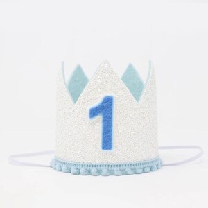 May include: White felt crown with blue felt accents and a blue felt number one. The crown has a white elastic band and a blue pom-pom trim.