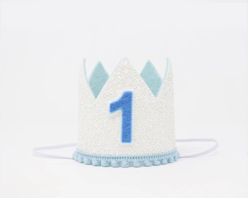 May include: White felt crown with blue felt accents and a blue felt number one. The crown has a white elastic band and a blue pom-pom trim.