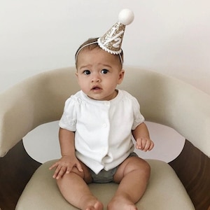 May include: A baby wearing a white shirt and gray shorts sits in a chair. The baby is wearing a gold glitter party hat with the number "1/2" on it.