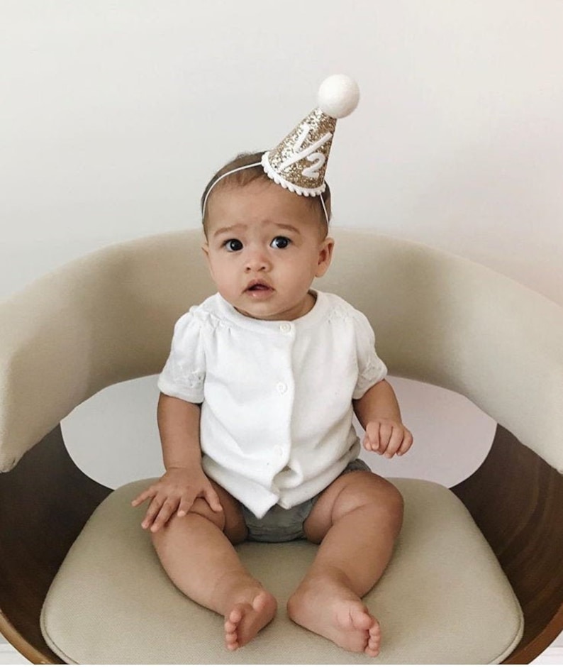 May include: A baby wearing a white shirt and gray shorts sits in a chair. The baby is wearing a gold glitter party hat with the number "1/2" on it.