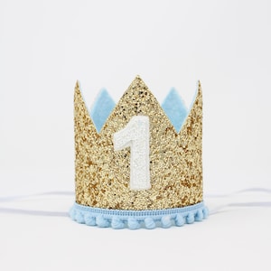 Puede incluir: Una corona dorada con purpurina, detalles en azul claro y el n&uacute;mero 1 blanco. La corona tiene un ribete de pomp&oacute;n azul claro y una banda el&aacute;stica. Ideal para un primer cumplea&ntilde;os.