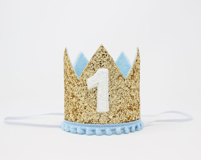 Puede incluir: Una corona dorada con purpurina, detalles en azul claro y el n&uacute;mero 1 blanco. La corona tiene un ribete de pomp&oacute;n azul claro y una banda el&aacute;stica. Ideal para un primer cumplea&ntilde;os.