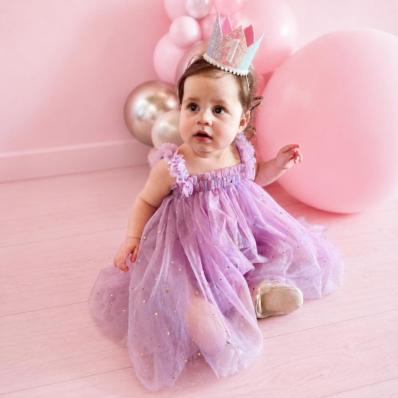 May include: A lavender tulle dress with gold star accents and ruffled shoulder straps, paired with a glittery crown featuring the number one. The dress has a layered, flowing skirt and a gathered bodice, creating a soft, textured appearance.
