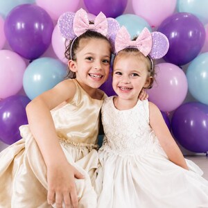 May include: Two headbands featuring light purple sequined ears and a large, shimmering light pink bow. The headbands are worn by two children, one in a gold satin dress and the other in a white lace dress, positioned in front of purple, pink, and blue balloons.