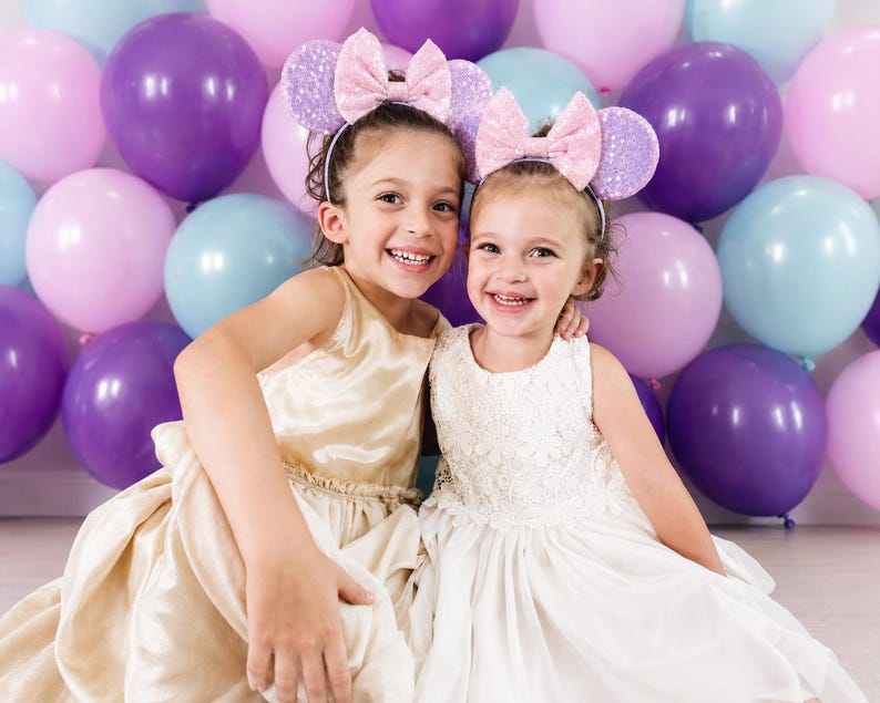 May include: Two headbands featuring light purple sequined ears and a large, shimmering light pink bow. The headbands are worn by two children, one in a gold satin dress and the other in a white lace dress, positioned in front of purple, pink, and blue balloons.