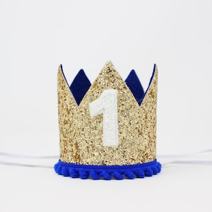 Puede incluir: Una corona dorada con purpurina y un n&uacute;mero 1 blanco aplicado. La corona tiene un ribete azul y detalles de fieltro azul. Una banda el&aacute;stica blanca est&aacute; unida a la corona.