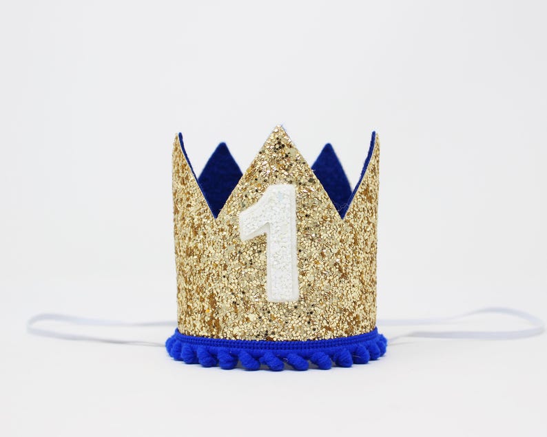 Puede incluir: Una corona dorada con purpurina y un n&uacute;mero 1 blanco aplicado. La corona tiene un ribete azul y detalles de fieltro azul. Una banda el&aacute;stica blanca est&aacute; unida a la corona.