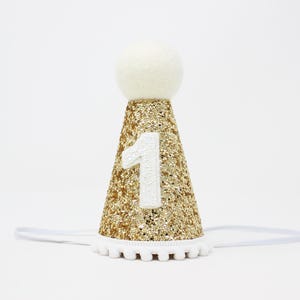May include: Gold glitter party hat with a white pom-pom trim and a white felt ball on top. The hat has a white felt number "1" on the front.