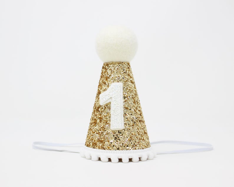 May include: Gold glitter party hat with a white pom-pom trim and a white felt ball on top. The hat has a white felt number "1" on the front.