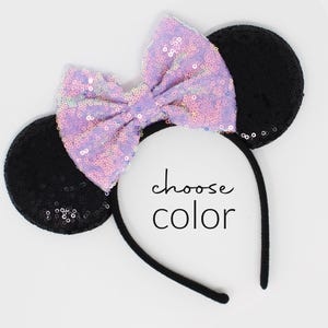 May include: A headband featuring two round black sequined ears and a large, central lavender sequined bow. The text "choose color" is printed in black in the center of the headband. The headband frame is thin and black.