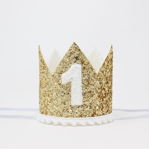 Puede incluir: Una corona dorada con purpurina y el n&uacute;mero "1" en blanco. La corona tiene un ribete de pomp&oacute;n blanco y una banda el&aacute;stica blanca. La corona est&aacute; dise&ntilde;ada para una celebraci&oacute;n de primer cumplea&ntilde;os.