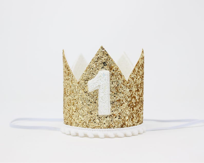 Puede incluir: Una corona dorada con purpurina y el n&uacute;mero "1" en blanco. La corona tiene un ribete de pomp&oacute;n blanco y una banda el&aacute;stica blanca. La corona est&aacute; dise&ntilde;ada para una celebraci&oacute;n de primer cumplea&ntilde;os.