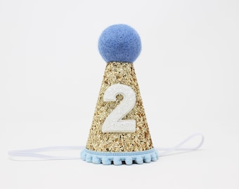 May include: A conical party hat covered in gold glitter with a white felt number 2 attached to the front. The hat is topped with a light blue felt pom-pom and features a light blue pom-pom trim along the bottom edge, with a thin white elastic strap.