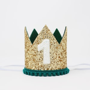 Puede incluir: Una corona dorada con purpurina y el n&uacute;mero 1 blanco. La corona tiene un interior verde oscuro y un ribete de pomp&oacute;n turquesa. Se adjunta una cinta blanca para asegurar la corona.