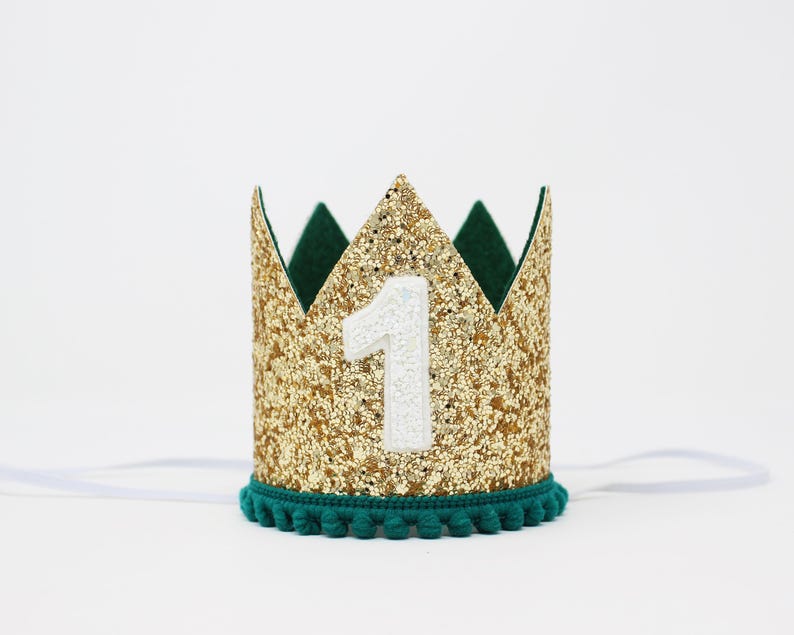 Puede incluir: Una corona dorada con purpurina y el n&uacute;mero 1 blanco. La corona tiene un interior verde oscuro y un ribete de pomp&oacute;n turquesa. Se adjunta una cinta blanca para asegurar la corona.