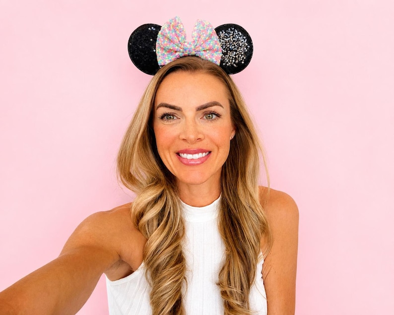 May include: A headband featuring two round black ears covered in dark glitter and a central bow with a multicolored pastel confetti pattern. The ears are positioned upright on a thin band, creating a distinct silhouette with a sparkling texture.