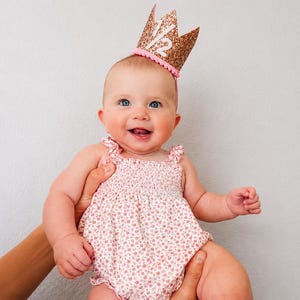 May include: A rose gold glitter crown with the white text 1/2 and a pink pom-pom trim, worn with a white romper featuring a small pink floral pattern, ruffled shoulder straps, and a smocked bodice design for a decorative appearance.