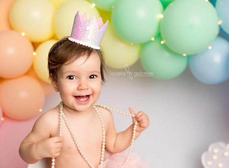 May include: A glittery pink and white crown with the number one on the front, featuring a white pom-pom trim along the base. The crown is worn on the head, and a long strand of white faux pearls is draped around the neck and shoulders.