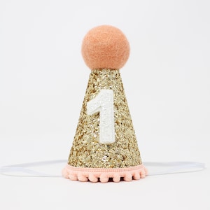 May include: Gold glitter party hat with a peach pom pom on top and a white felt number one on the front. The hat is attached to a clear elastic headband.