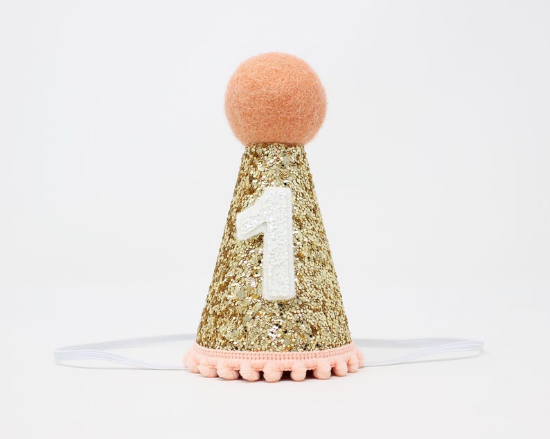 May include: Gold glitter party hat with a peach pom pom on top and a white felt number one on the front. The hat is attached to a clear elastic headband.