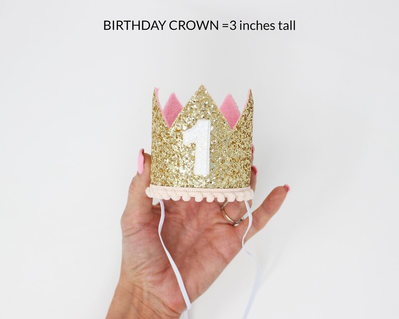 Puede incluir: Una corona de cumplea&ntilde;os dorada con purpurina, detalles rosas y el n&uacute;mero 1. La corona mide 7.6 cm de alto y tiene un ribete color crema con cintas blancas. El texto "BIRTHDAY CROWN = 3 inches tall" est&aacute; arriba.
