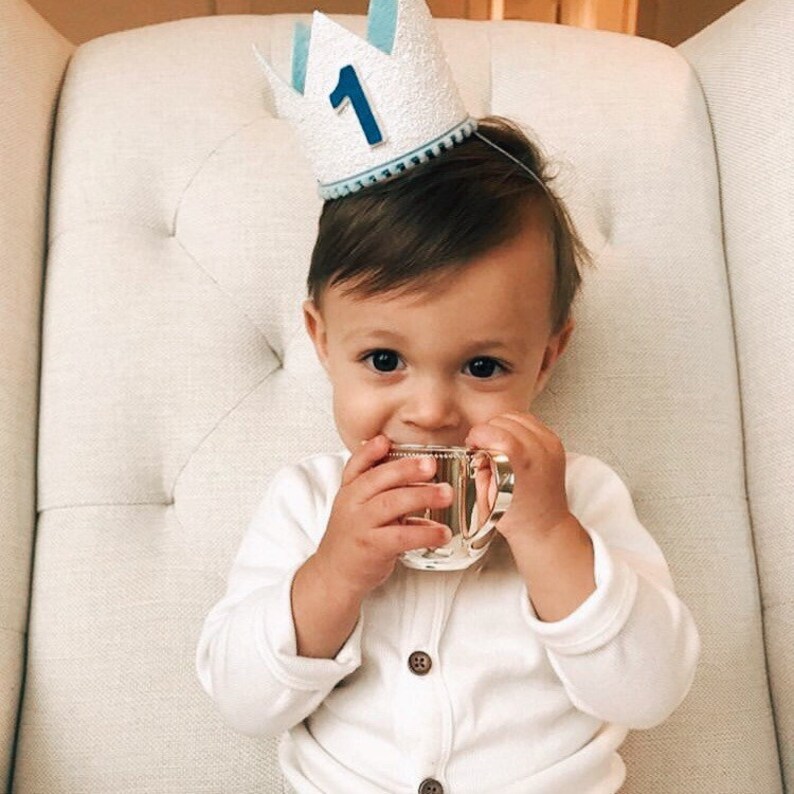 May include: A baby wearing a white and blue crown with the number "1" on it, sitting in a white chair and holding a small cup.
