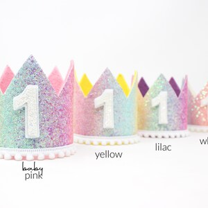 May include: Four glittery crowns with a white number 1 on the front, featuring pom-pom trim at the base. The crowns are labeled baby pink, yellow, lilac, and white, showcasing a gradient of pastel colors with a sparkling texture and pointed tops.
