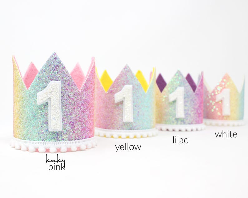 May include: Four glittery crowns with a white number 1 on the front, featuring pom-pom trim at the base. The crowns are labeled baby pink, yellow, lilac, and white, showcasing a gradient of pastel colors with a sparkling texture and pointed tops.