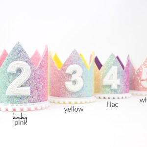 May include: Four glittery rainbow-colored crowns with scalloped edges, each featuring a large white felt number from two to five on the front. The base of each crown is trimmed with a white pom-pom fringe. Text labels include baby pink, yellow, lilac, and white.