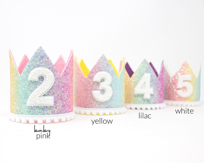 May include: Four glittery rainbow-colored crowns with scalloped edges, each featuring a large white felt number from two to five on the front. The base of each crown is trimmed with a white pom-pom fringe. Text labels include baby pink, yellow, lilac, and white.