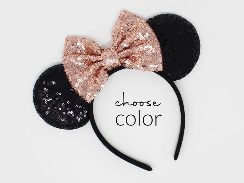 May include: A headband featuring two round black sequined ears and a large rose gold sequined bow centered between them. The text "choose color" is printed in black in the center of the headband, which has a plain black band.