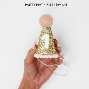 May include: Gold glitter party hat with a white number one and pink pom pom trim. The hat is 3.5 inches tall.