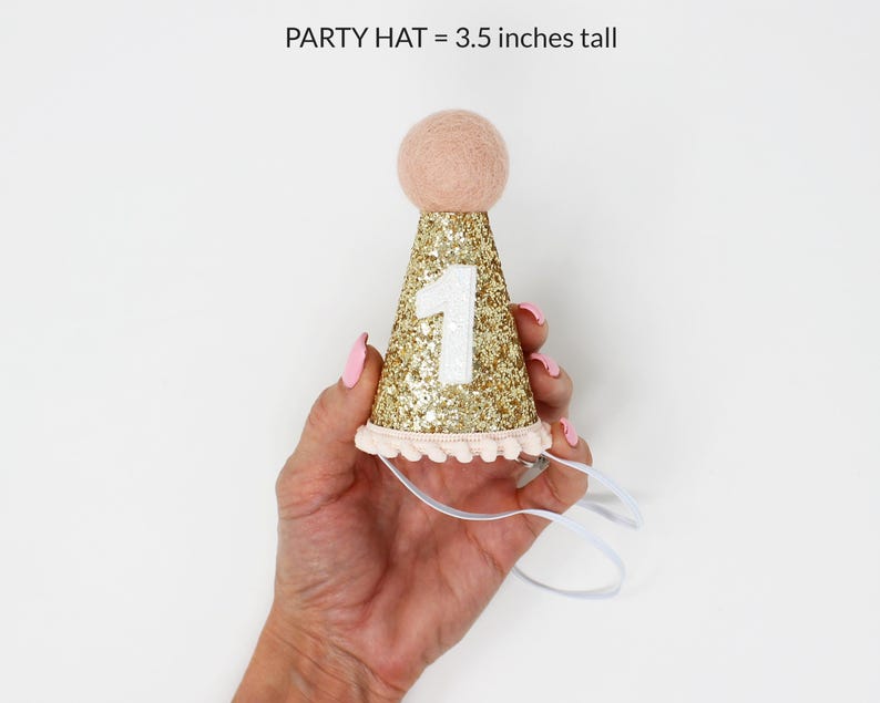 May include: Gold glitter party hat with a white number one and pink pom pom trim. The hat is 3.5 inches tall.