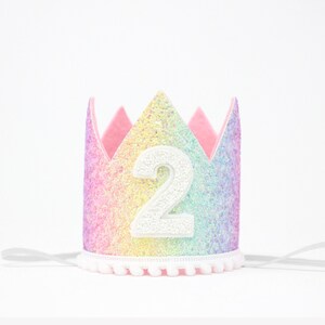 May include: A crown featuring a rainbow glitter finish with a white number 2 centered on the front. The crown has a scalloped top edge, a white pom-pom trim along the bottom, and thin white elastic straps extending from each side for securing.