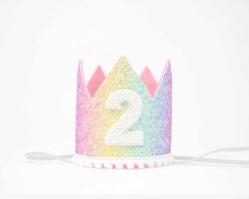 May include: A crown featuring a rainbow glitter finish with a white number 2 centered on the front. The crown has a scalloped top edge, a white pom-pom trim along the bottom, and thin white elastic straps extending from each side for securing.