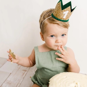 Puede incluir: Un ni&ntilde;o peque&ntilde;o con una corona dorada con purpurina, ribete verde y el n&uacute;mero 1. El ni&ntilde;o lleva un pelele verde y sostiene un objeto dorado. Una tarta blanca est&aacute; en primer plano.