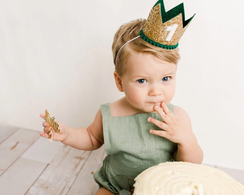 Puede incluir: Un ni&ntilde;o peque&ntilde;o con una corona dorada con purpurina, ribete verde y el n&uacute;mero 1. El ni&ntilde;o lleva un pelele verde y sostiene un objeto dorado. Una tarta blanca est&aacute; en primer plano.