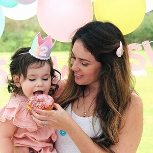 May include: A small child wearing a pastel rainbow-colored felt crown with the number 2 on the front and a ruffled dusty pink dress, eating a pink frosted donut with colorful sprinkles. The child is held by an adult wearing a white tank top.
