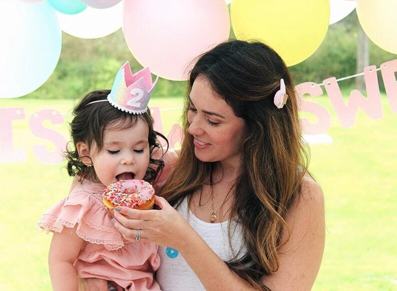 May include: A small child wearing a pastel rainbow-colored felt crown with the number 2 on the front and a ruffled dusty pink dress, eating a pink frosted donut with colorful sprinkles. The child is held by an adult wearing a white tank top.