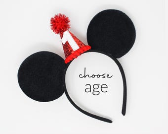 May include: A black headband featuring two large round black ears and a small red glittery party hat attached to the top. The hat is topped with a red pom-pom and displays a white number one. The text choose age is printed in the center.