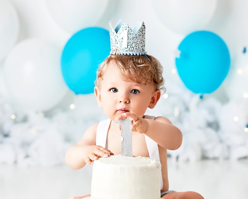 Puede incluir: Un beb&eacute; con una corona de purpurina plateada y sosteniendo un adorno de pastel con el n&uacute;mero uno plateado. El beb&eacute; est&aacute; frente a un pastel blanco y globos blancos y azules.
