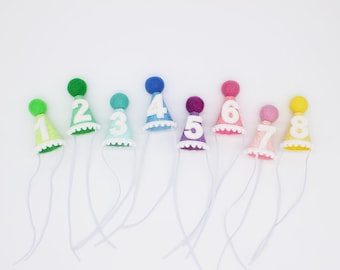 May include: A set of eight miniature cone-shaped party hats in various colors, each topped with a matching felt pom-pom. Each hat features a large white felt number from one to eight, a white scalloped trim at the base, and a thin white elastic chin strap.