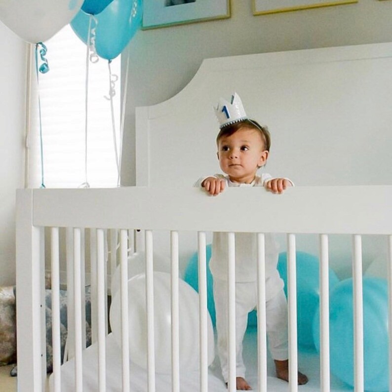 May include: A baby wearing a white shirt and a blue and white crown with the number "1" on it, standing in a white crib. The crib has white slats and a white mattress. There are blue and white balloons in the crib.