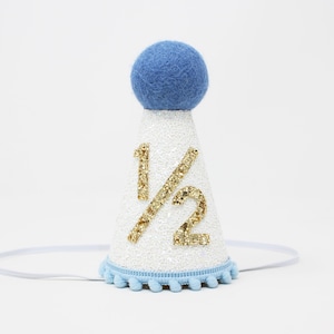 May include: A white cone-shaped party hat with gold glitter "1/2" text. The hat has a blue pom-pom on top and a blue trim with small pom-poms at the base. A thin white elastic band is attached.