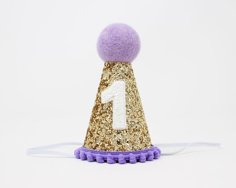 May include: A purple and gold glitter party hat with a white felt number one on the front. The hat has a purple pom-pom on top and a white elastic band.
