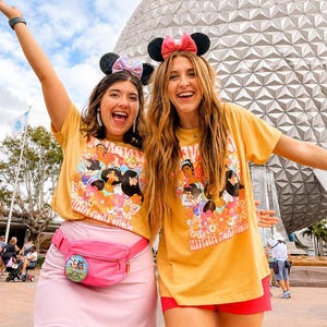 May include: Two mustard yellow t-shirts featuring a colorful graphic print of various animated princesses surrounded by orange and pink flowers. One shirt is paired with a light pink skirt and a bright pink fanny pack with a circular pin attached.