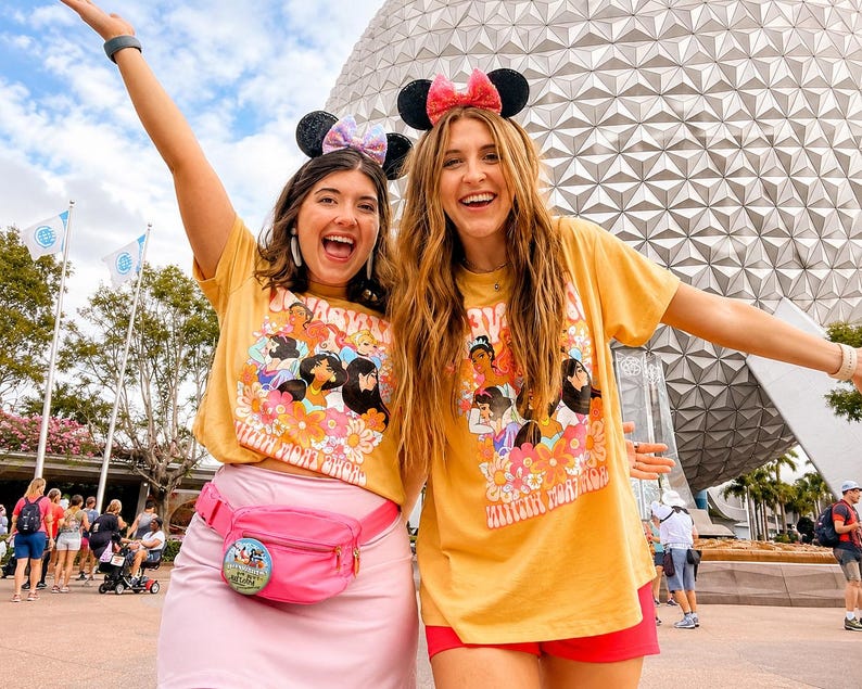 May include: Two mustard yellow t-shirts featuring a colorful graphic print of various animated princesses surrounded by orange and pink flowers. One shirt is paired with a light pink skirt and a bright pink fanny pack with a circular pin attached.