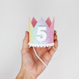 May include: A crown-shaped party hat featuring a pastel rainbow glitter finish and a white number five centered on the front. The base is trimmed with a white pom-pom fringe and includes a thin white elastic chin strap for secure placement.