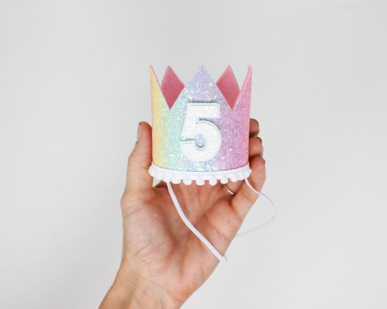 May include: A crown-shaped party hat featuring a pastel rainbow glitter finish and a white number five centered on the front. The base is trimmed with a white pom-pom fringe and includes a thin white elastic chin strap for secure placement.